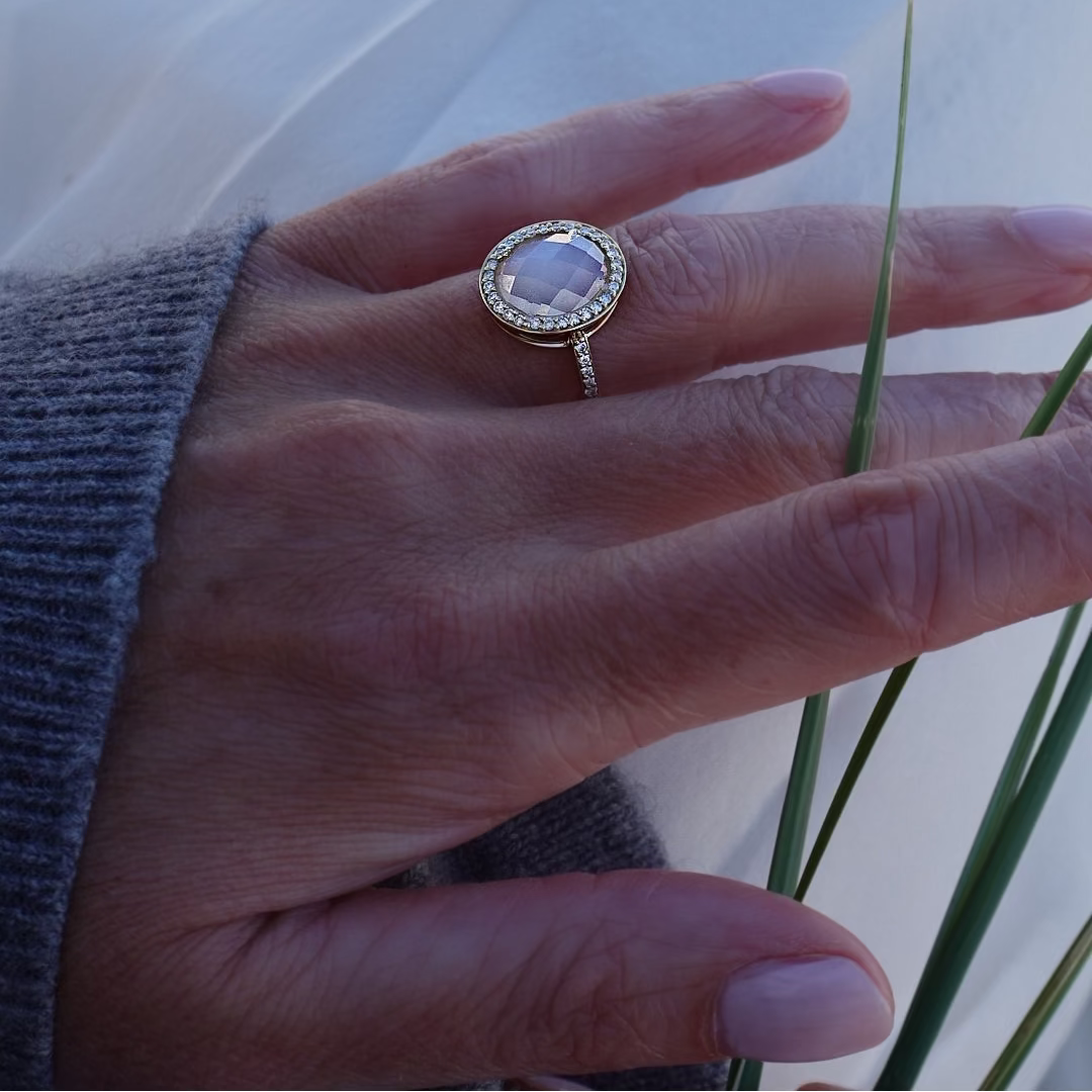 A gold ring with a round clear gemstone and a halo of small clear stones, worn on a person's finger. The hand is resting against a white fabric background with some green grass blades visible.