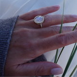 A gold ring with a round clear gemstone and a halo of small clear stones, worn on a person's finger. The hand is resting against a white fabric background with some green grass blades visible.