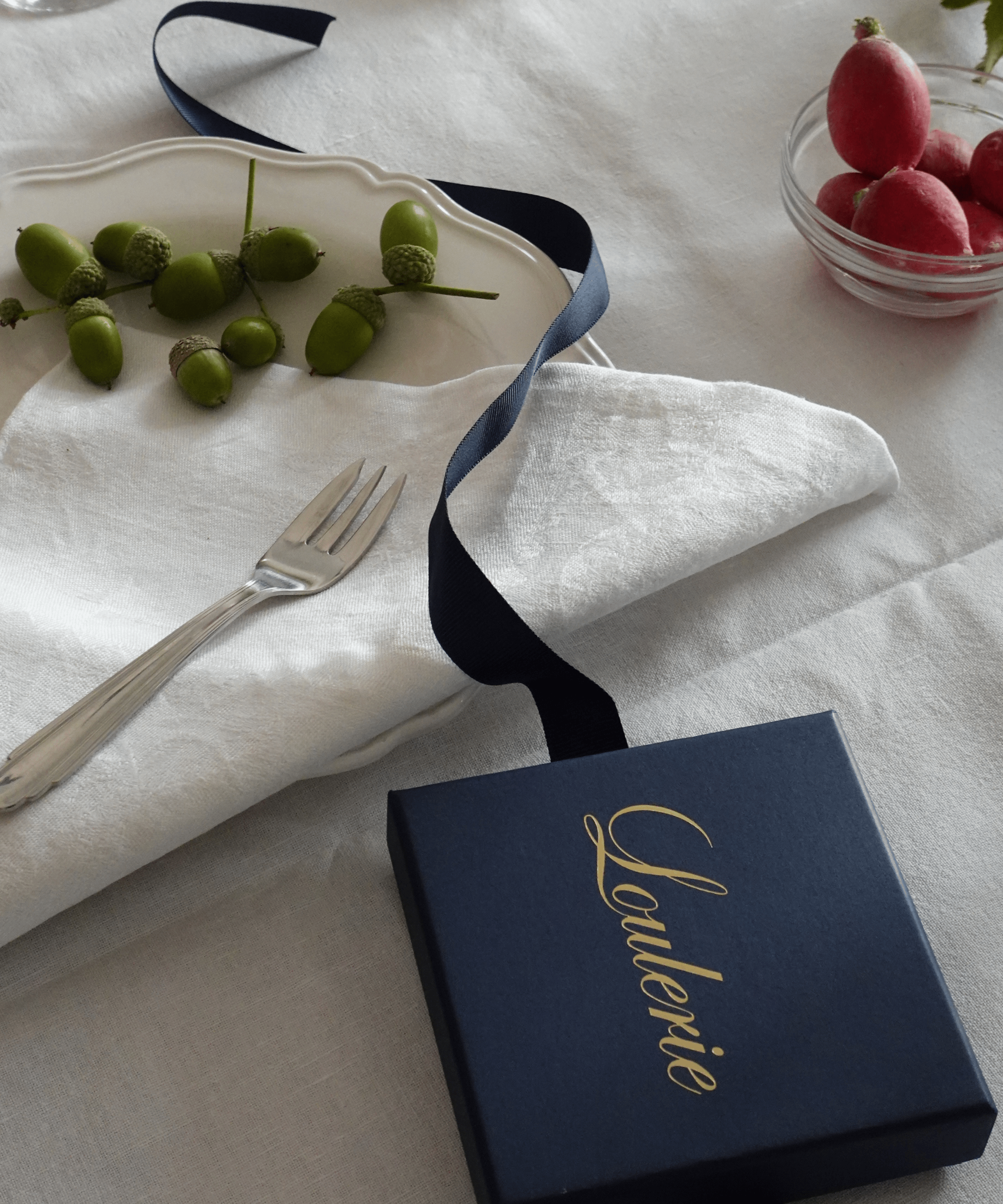 A navy blue gift box with elegant gold foil script reading 'Loulerie' on the lid, styled on a white linen tablecloth with a white napkin, silver fork, green acorns, and a small bowl of red fruit.