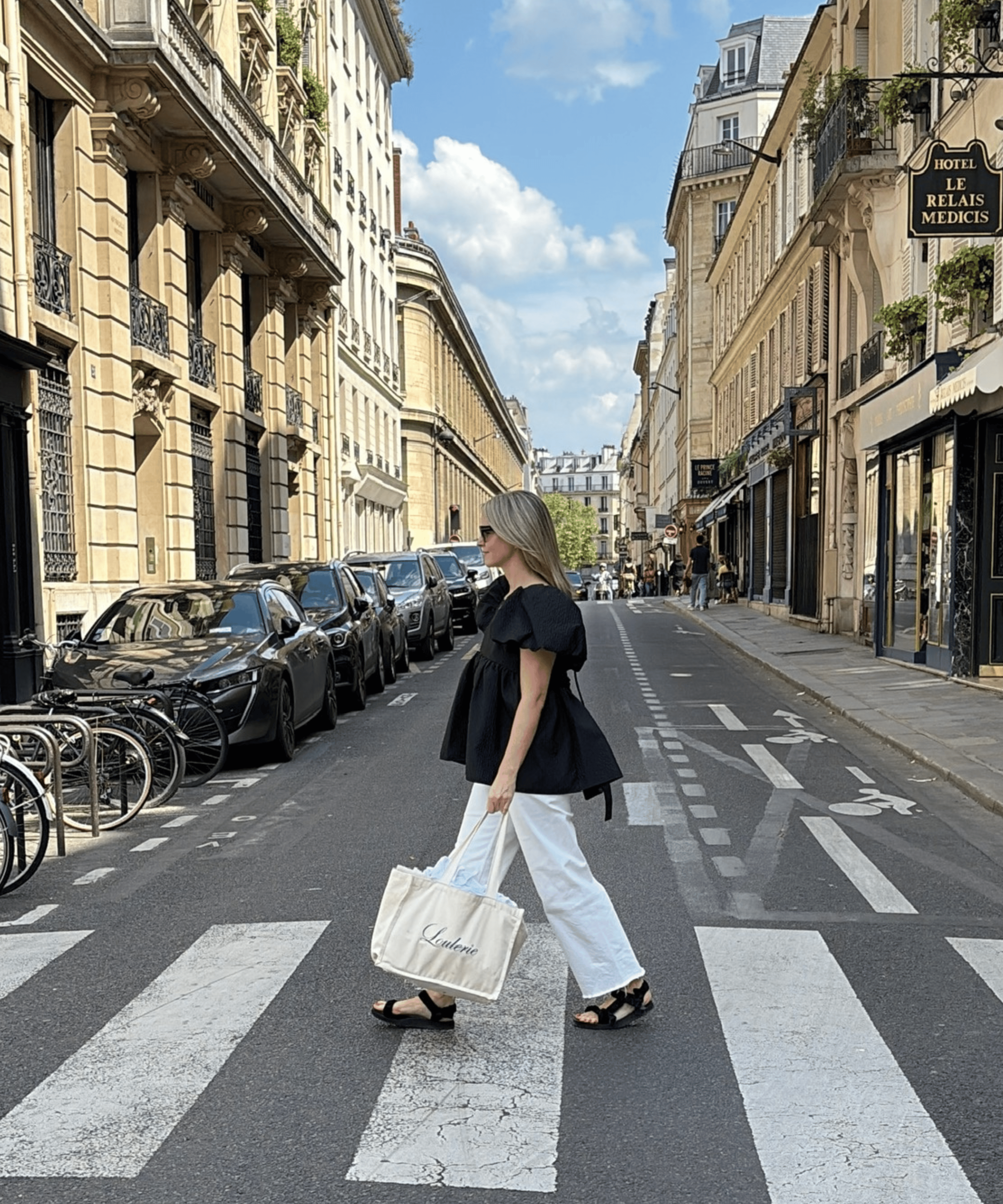 A woman walks across a crosswalk on a Parisian street, wearing a black puff-sleeve top, white wide-leg pants, and black sandals, while carrying a large white  Loulerie-branded tote bag.