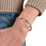 A close-up of a person's wrist wearing a gold and silver pavé snake cuff bracelet, styled with a beige ribbed sweater and blue jeans against a white background.