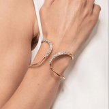 A close-up of a person's wrist and hand wearing a pair of open cuff bracelets. The bracelets are gold-toned with pavé-set clear stones along the curved front edges, photographed against a soft white background.