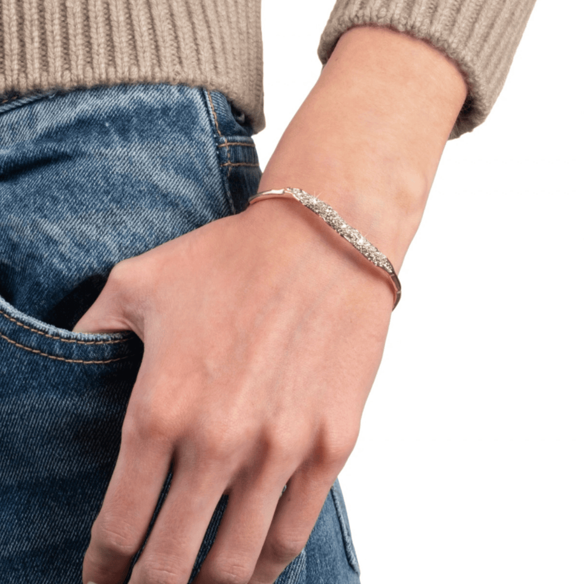 A close-up of a person's hand and wrist wearing a slim gold bangle bracelet with a row of clear pavé-set stones, paired with blue jeans and a beige ribbed sweater.