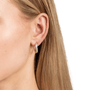 A close-up of a woman's ear wearing a small teardrop-shaped drop earring with a textured rose gold setting and clear stones, against a plain white background.