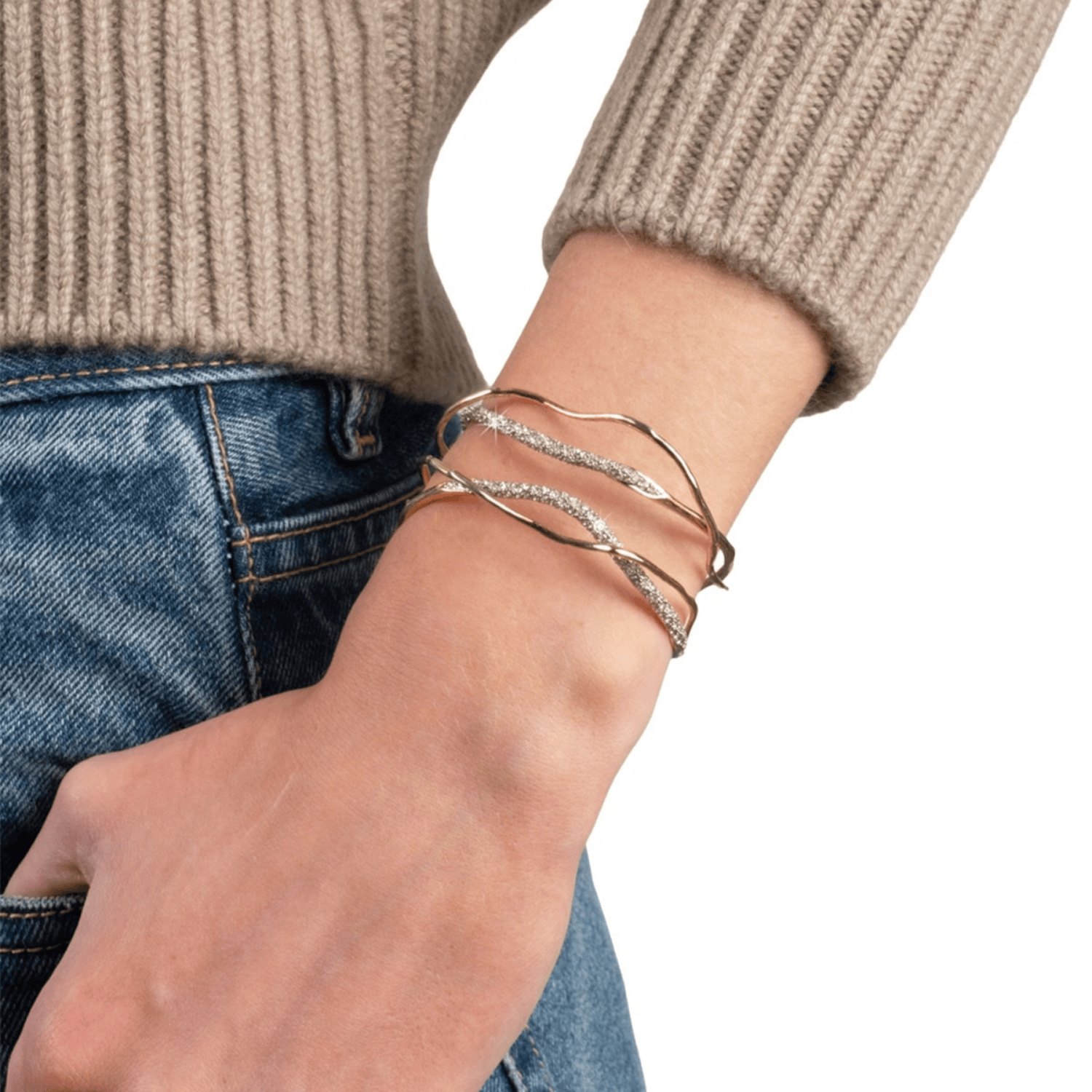 A close-up of a person's wrist wearing three twisted wrap bracelets, including two gold-toned and one silver-toned with clear stones, styled with a beige ribbed sweater and blue jeans.