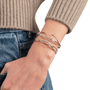 A close-up of a person's wrist wearing three twisted wrap bracelets, including two gold-toned and one silver-toned with clear stones, styled with a beige ribbed sweater and blue jeans.