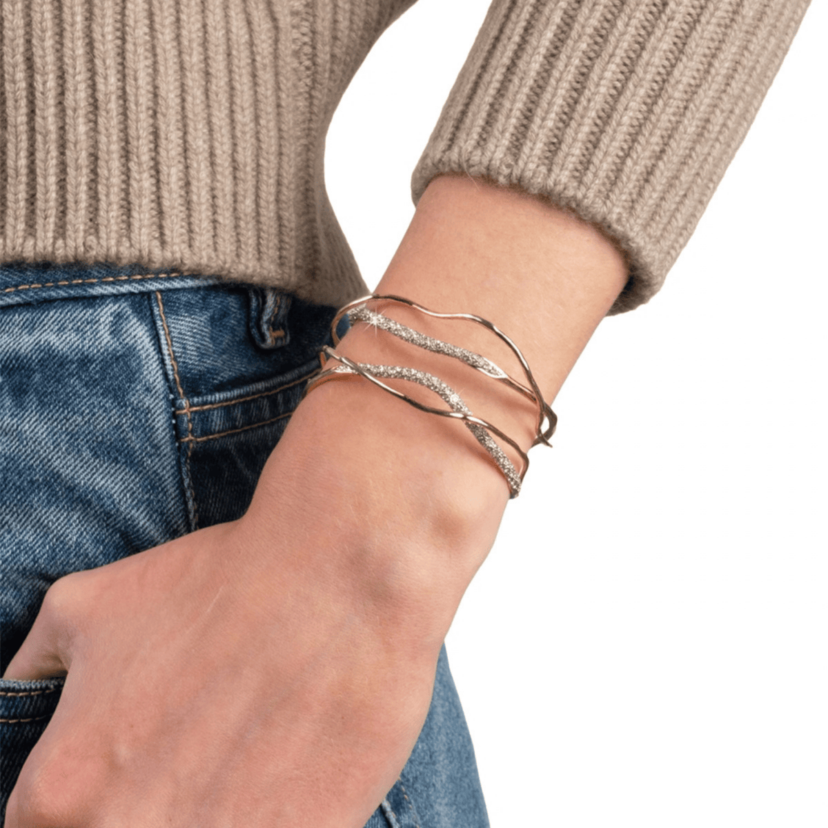 A close-up of a person's wrist wearing three twisted wrap bracelets, including two gold-toned and one silver-toned with clear stones, styled with a beige ribbed sweater and blue jeans.