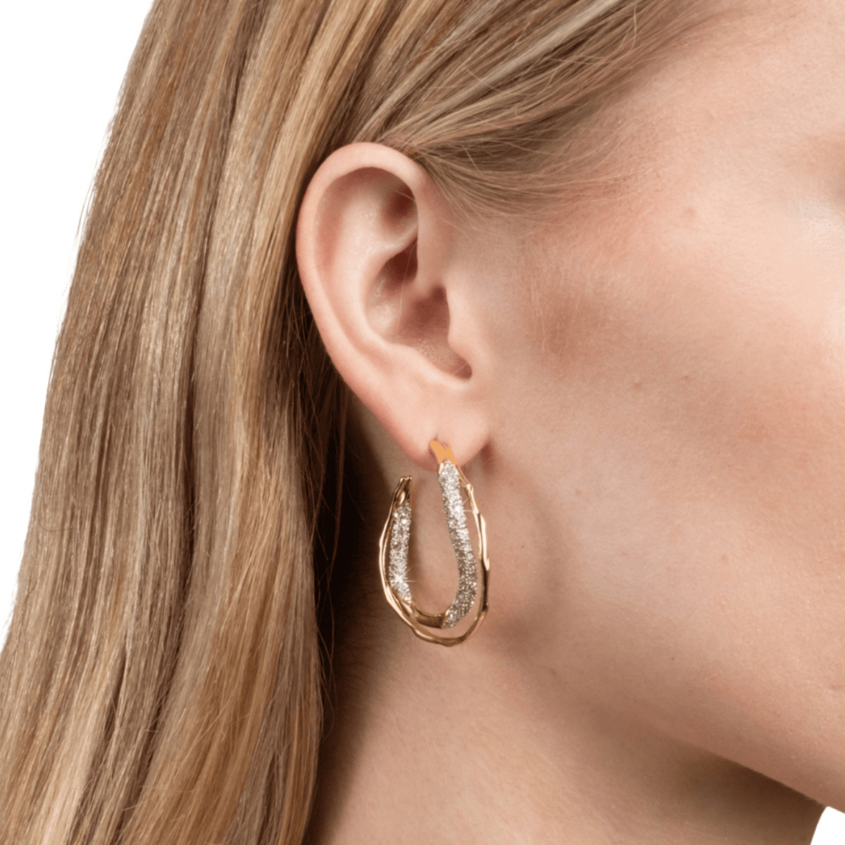 A close-up of a woman's ear wearing a gold hoop earring with a textured, diamond-like silver inlay, set against a clean white background.