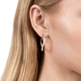 A close-up of a woman's ear wearing a gold crescent-shaped hoop earring set with small clear stones, against a clean white background.