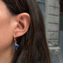 A close-up of a person's ear wearing a small gold hoop earring with a dangling irregular blue gemstone.