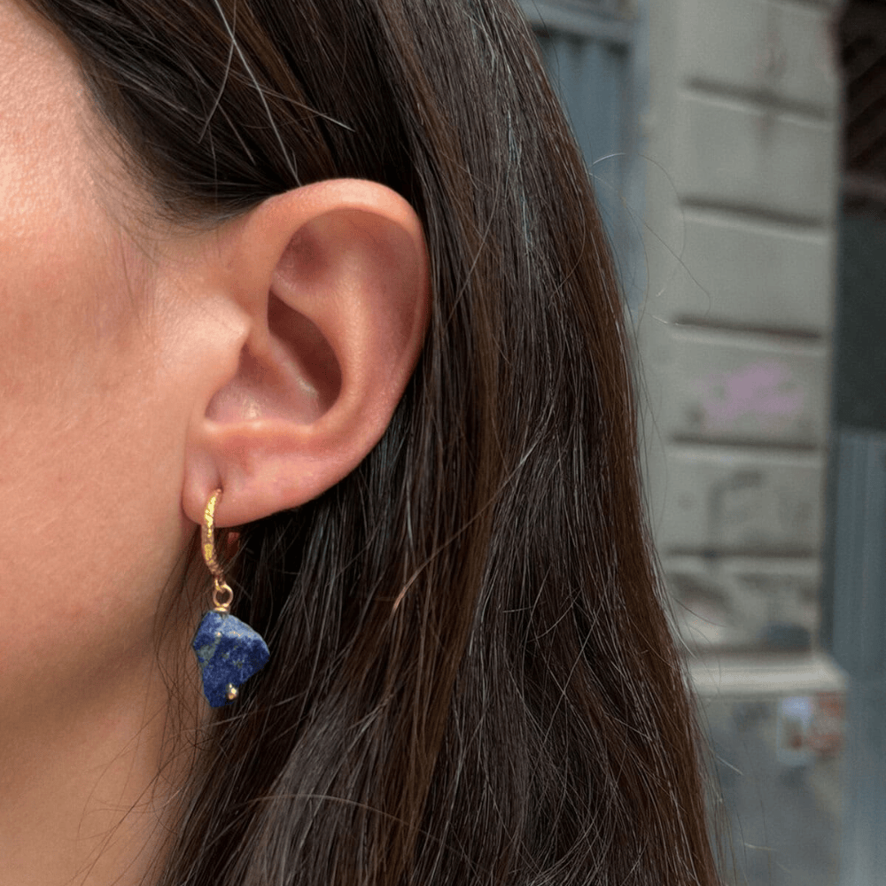A close-up of a person's ear wearing a small gold hoop earring with a dangling irregular blue gemstone.