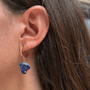 A close-up of a person's ear wearing a gold huggie hoop earring with a dangling natural blue lapis lazuli stone.