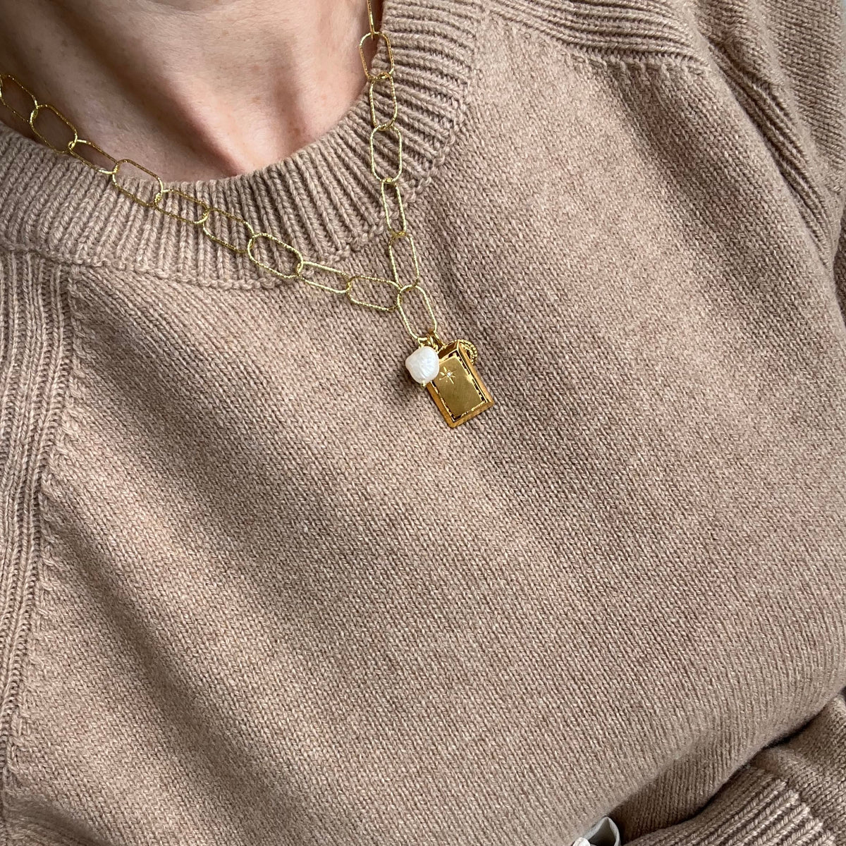 A close-up of a person wearing a gold chain necklace with a small rectangular gold pendant and a white bead, styled over a beige knit sweater.
