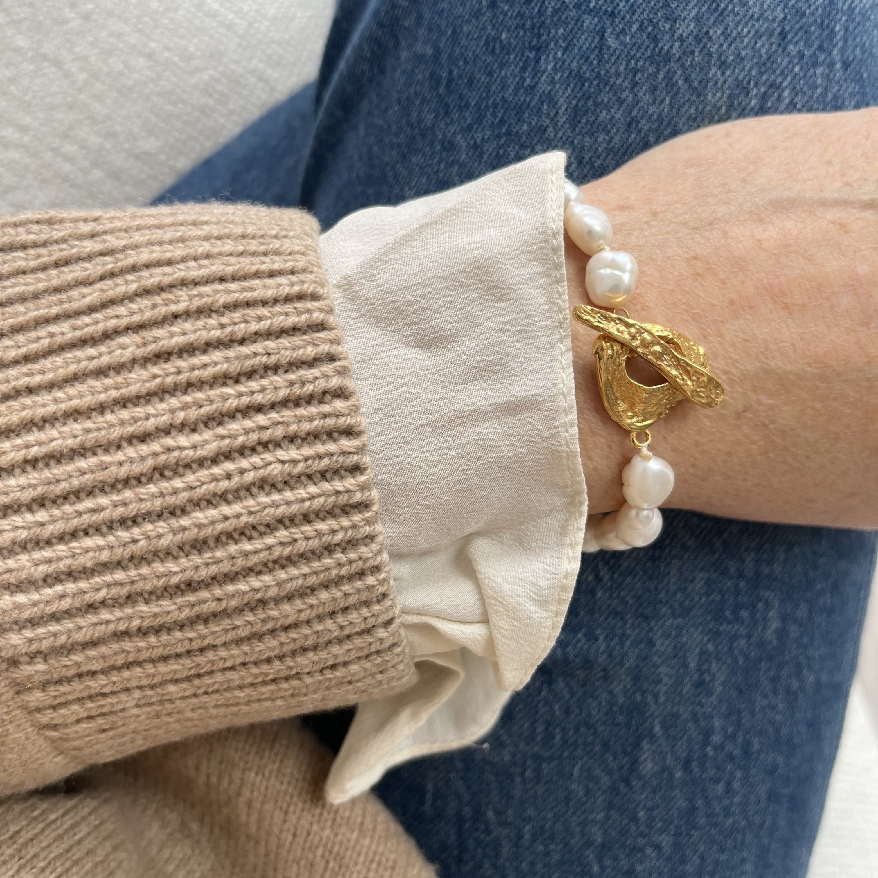 A close-up of a person's wrist wearing a gold and white pearl bracelet, styled with a cream ruffled shirt cuff, a beige ribbed sweater, and blue jeans.
