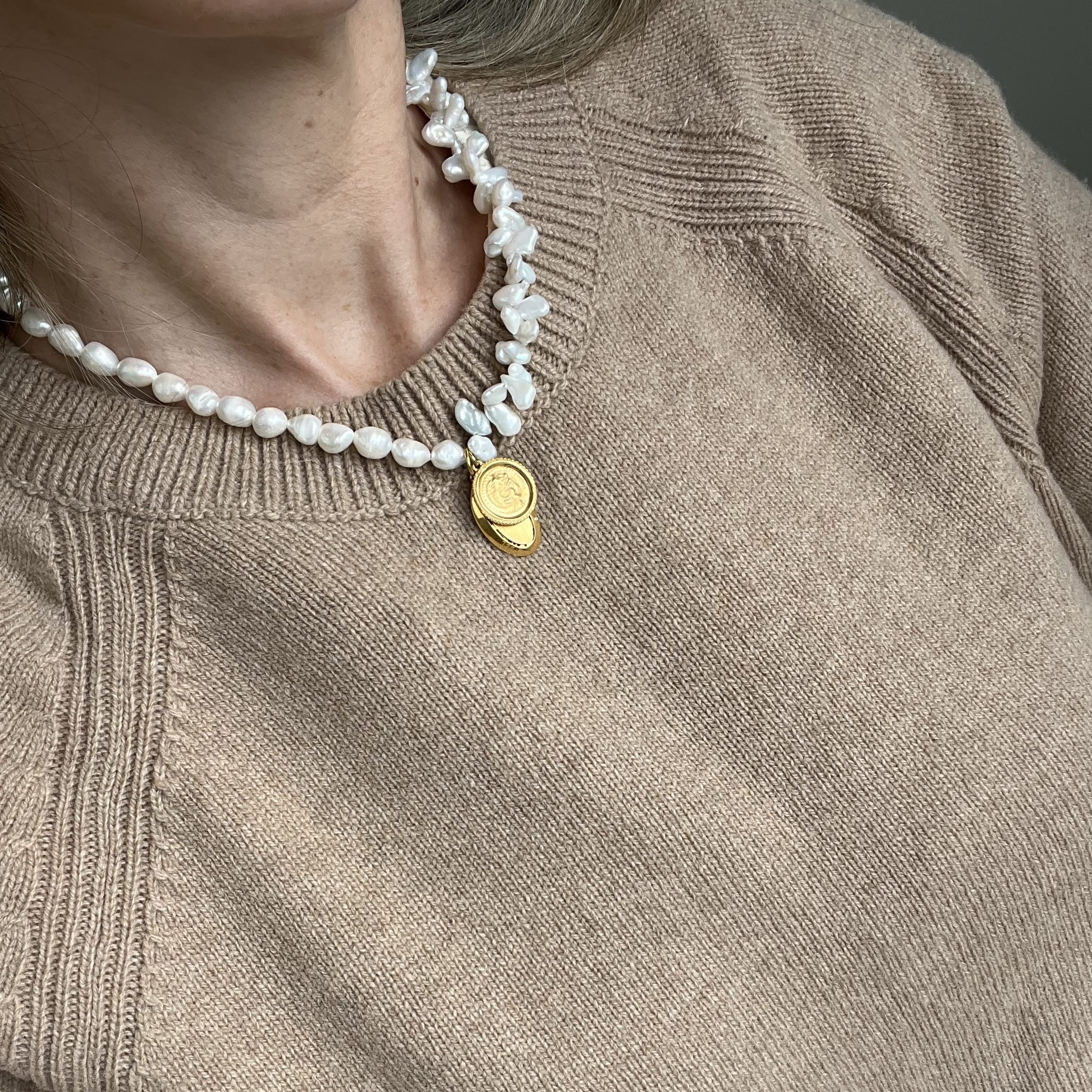 A close-up of a person wearing a chunky white baroque pearl necklace with a gold-toned oval pendant, paired with a beige knit sweater.