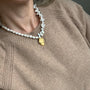 A close-up of a person wearing a chunky white baroque pearl necklace with a gold-toned oval pendant, paired with a beige knit sweater.