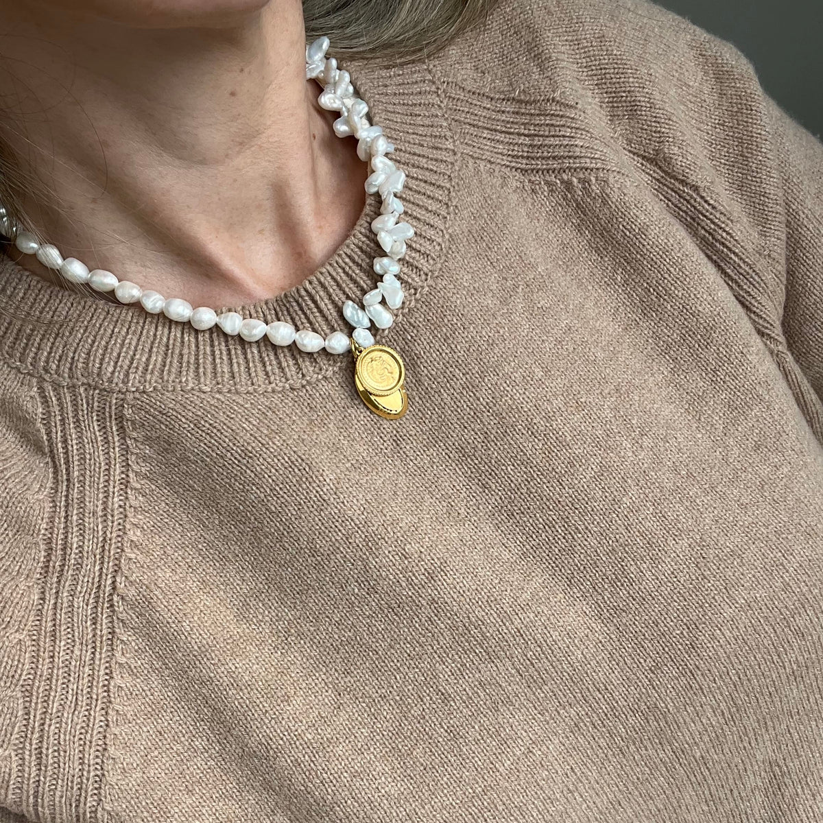 A close-up of a person wearing a chunky white baroque pearl necklace with a gold-toned oval pendant, paired with a beige knit sweater.