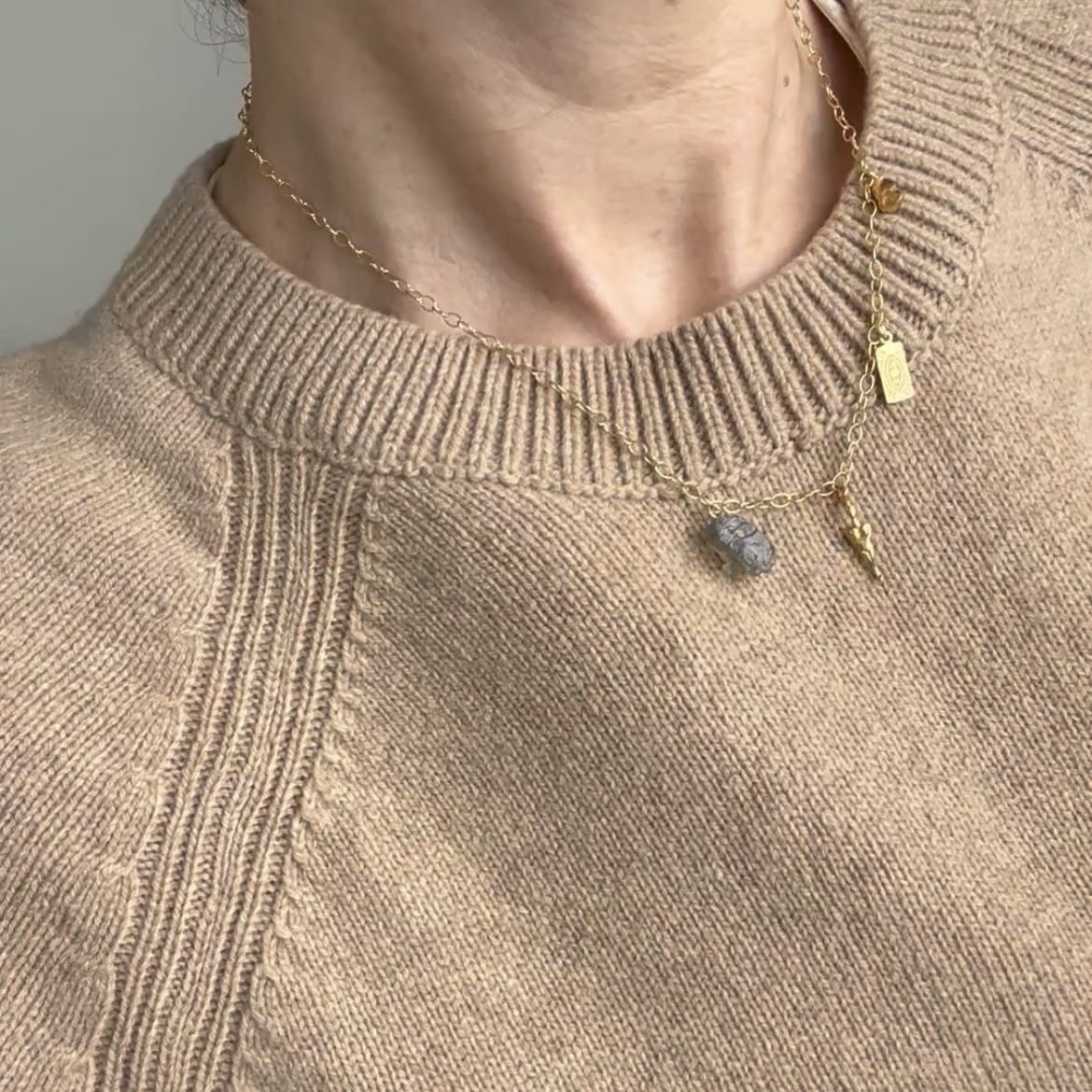 A close-up of a person wearing a delicate gold necklace with a small gray stone pendant and a tiny gold charm, styled over a beige knit sweater.