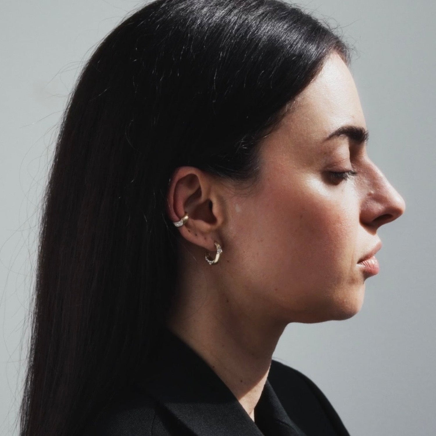 A close-up side profile of a woman with long dark hair wearing small gold ear piercings, including a hoop in her cartilage and a small hoop in her lobe, against a plain light gray background.