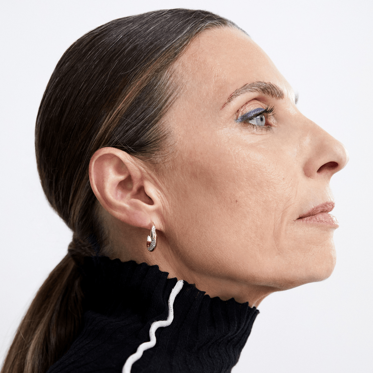 A close-up side profile of a woman with dark hair pulled back, wearing a small silver hoop earring with a clear gemstone, against a plain light background.