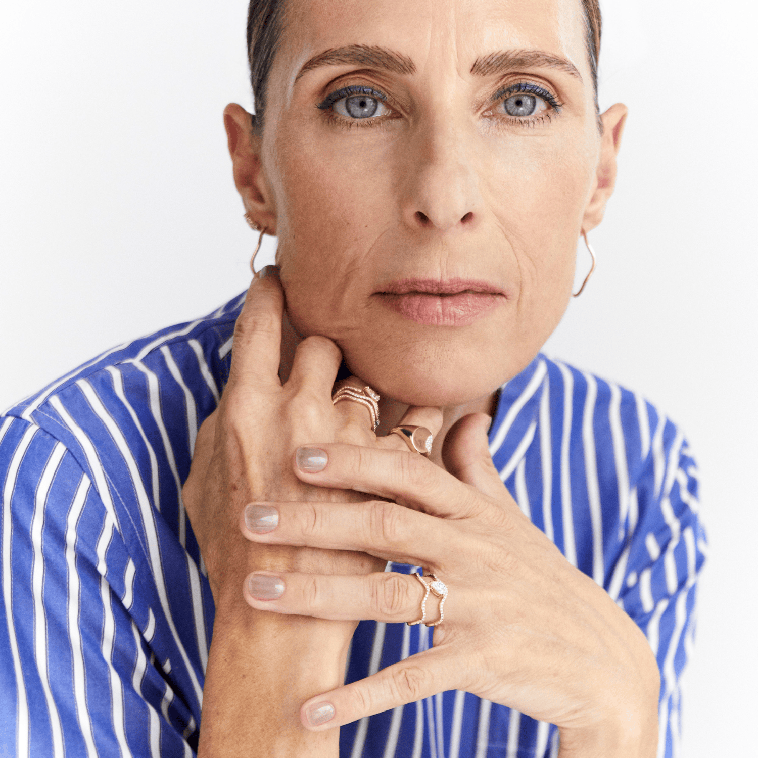 A close-up portrait of a woman with blue eyes and short hair, wearing a blue and white striped shirt and multiple gold rings, posed against a clean white background.