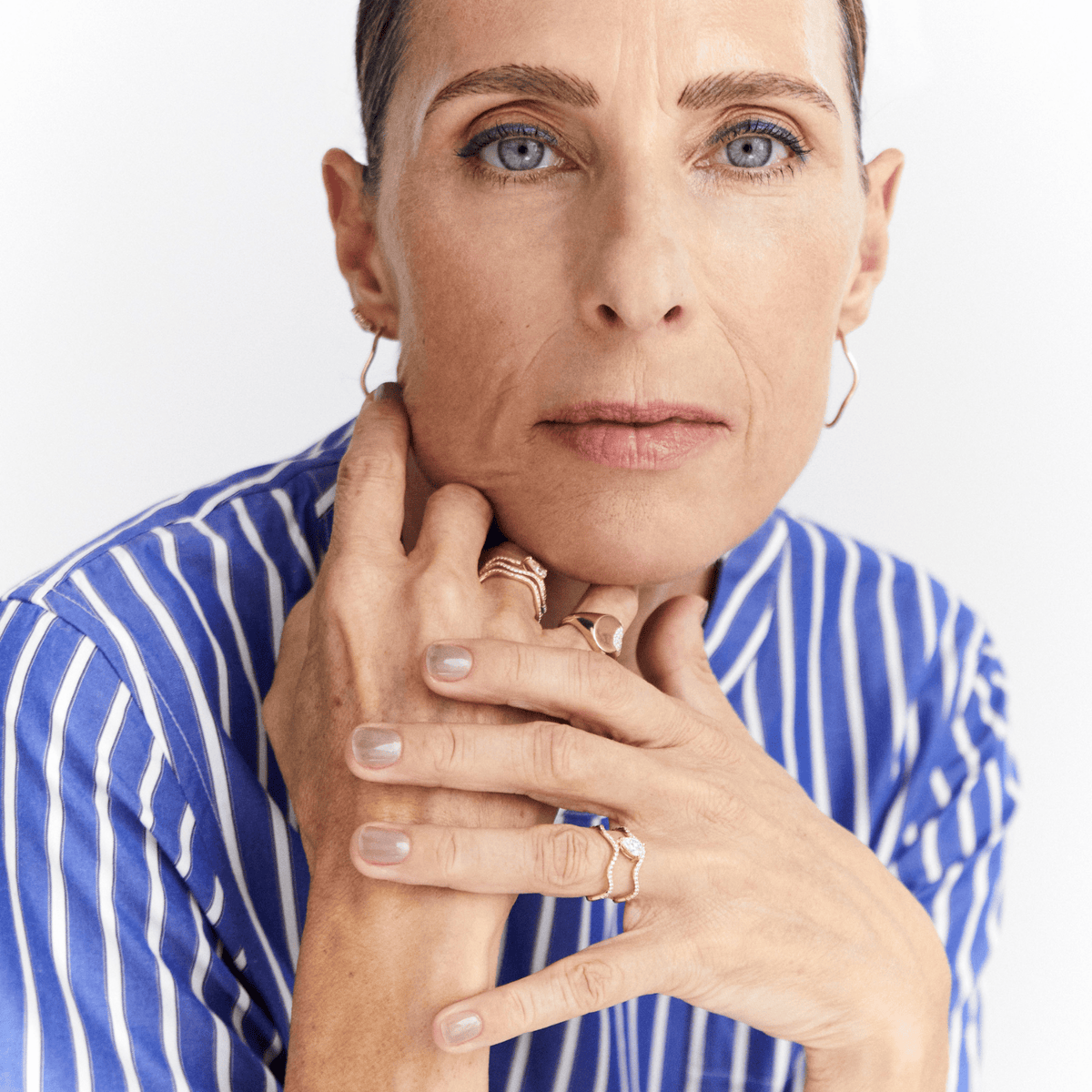 A close-up portrait of a woman with blue eyes and short hair, wearing a blue and white striped shirt and multiple gold rings, posed against a clean white background.