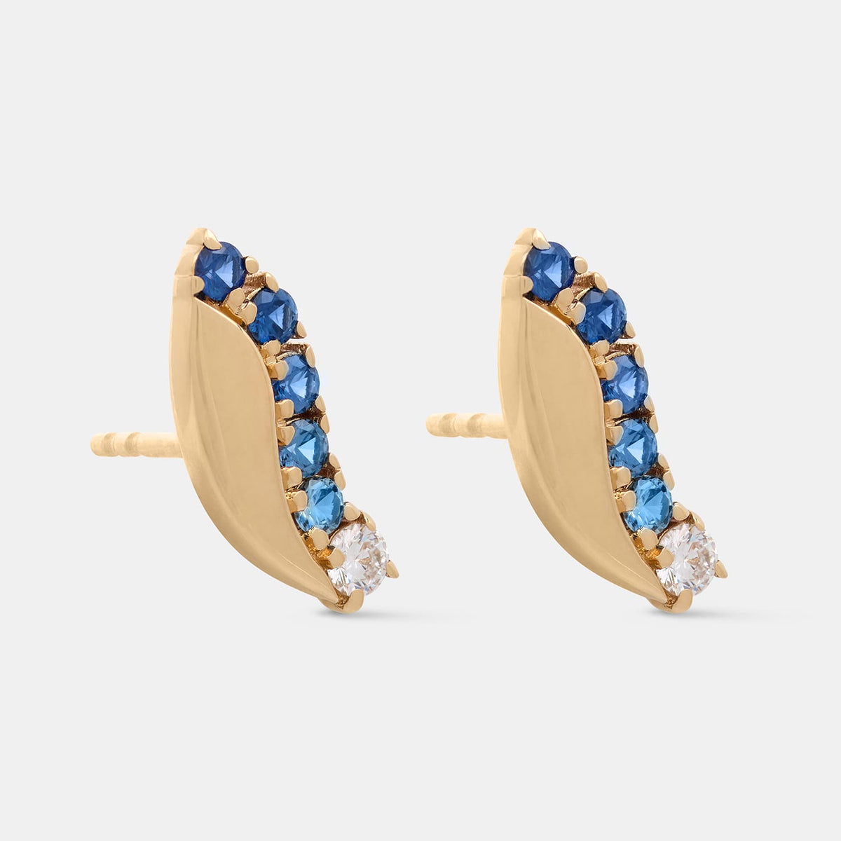 A pair of gold crescent-shaped stud earrings decorated with a row of faceted blue gemstones and a small clear stone at the tip, shown on a light gray background.