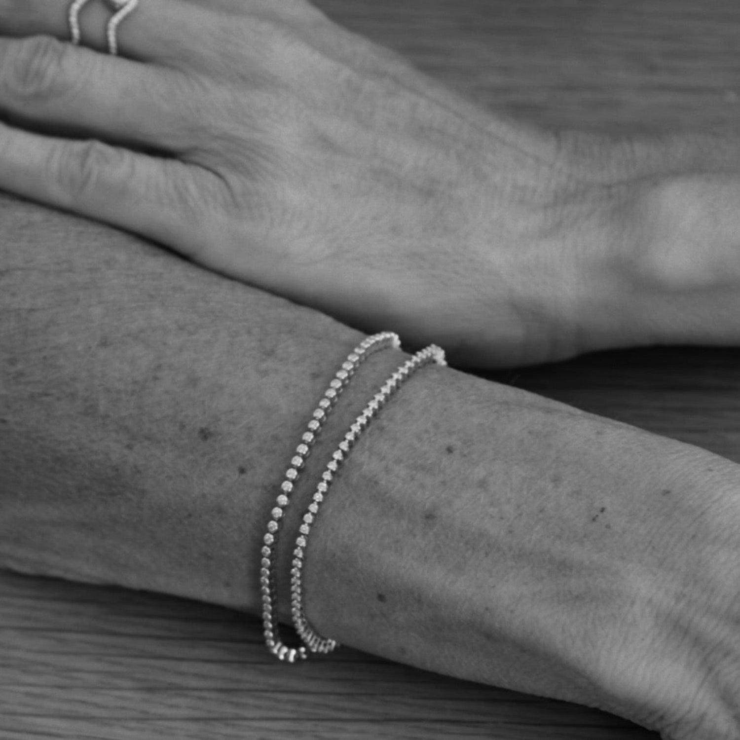 A close-up black and white photo of a person's wrist wearing two delicate chain bracelets, with one hand resting above the wrist on a wooden surface.