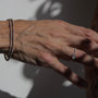 A close-up of a person's hand wearing a delicate silver ring on one finger and a beaded silver bracelet on the other wrist, set against a neutral background with dramatic shadows.