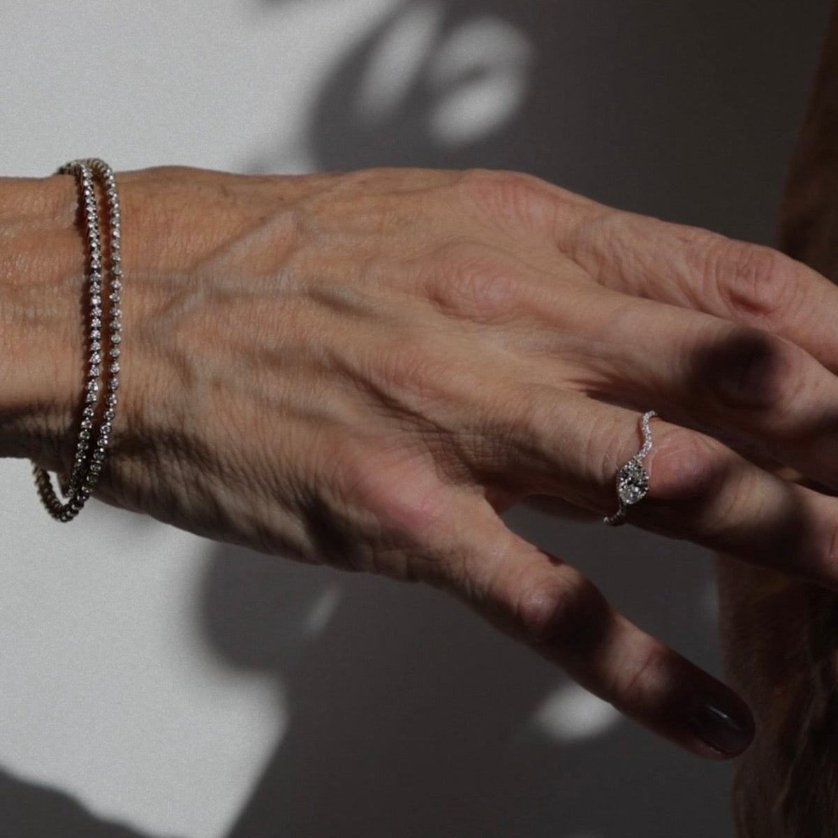 A close-up of a person's hand wearing a delicate silver ring on one finger and a beaded silver bracelet on the other wrist, set against a neutral background with dramatic shadows.