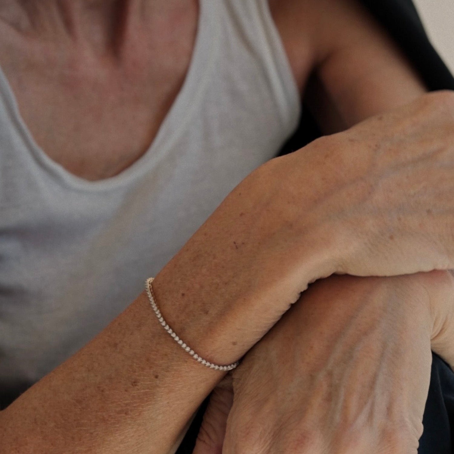 A close-up of a delicate silver beaded chain bracelet worn on a person's wrist, with their hands crossed over their lap and a white tank top visible in the background.
