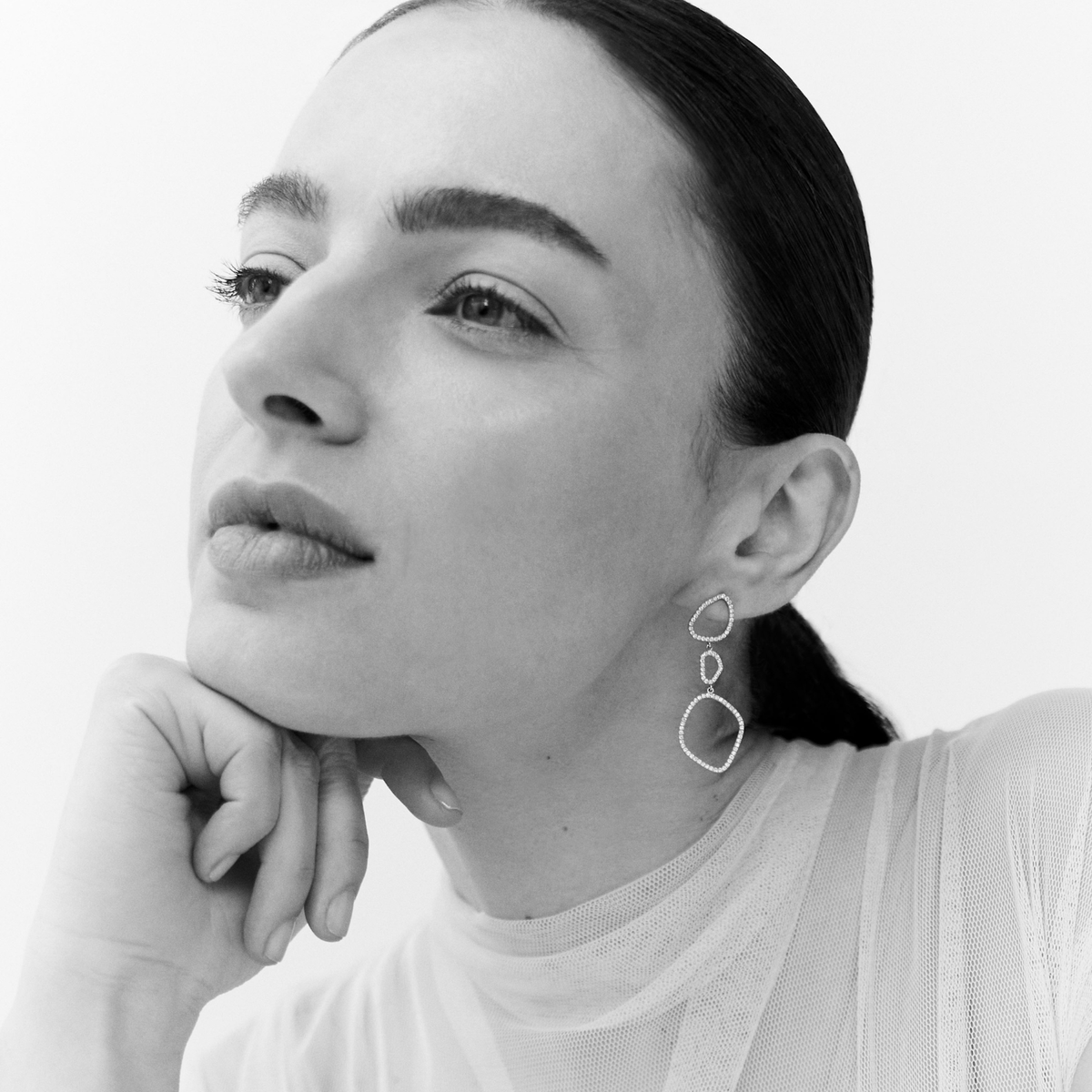 A close-up black and white portrait of a woman wearing a pair of dangling geometric wire earrings, with her hand resting under her chin against a plain light background.