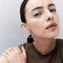 A close-up portrait of a woman wearing a thin, open circular hoop earring in silver, paired with a dark brown sheer top against a light gray background.