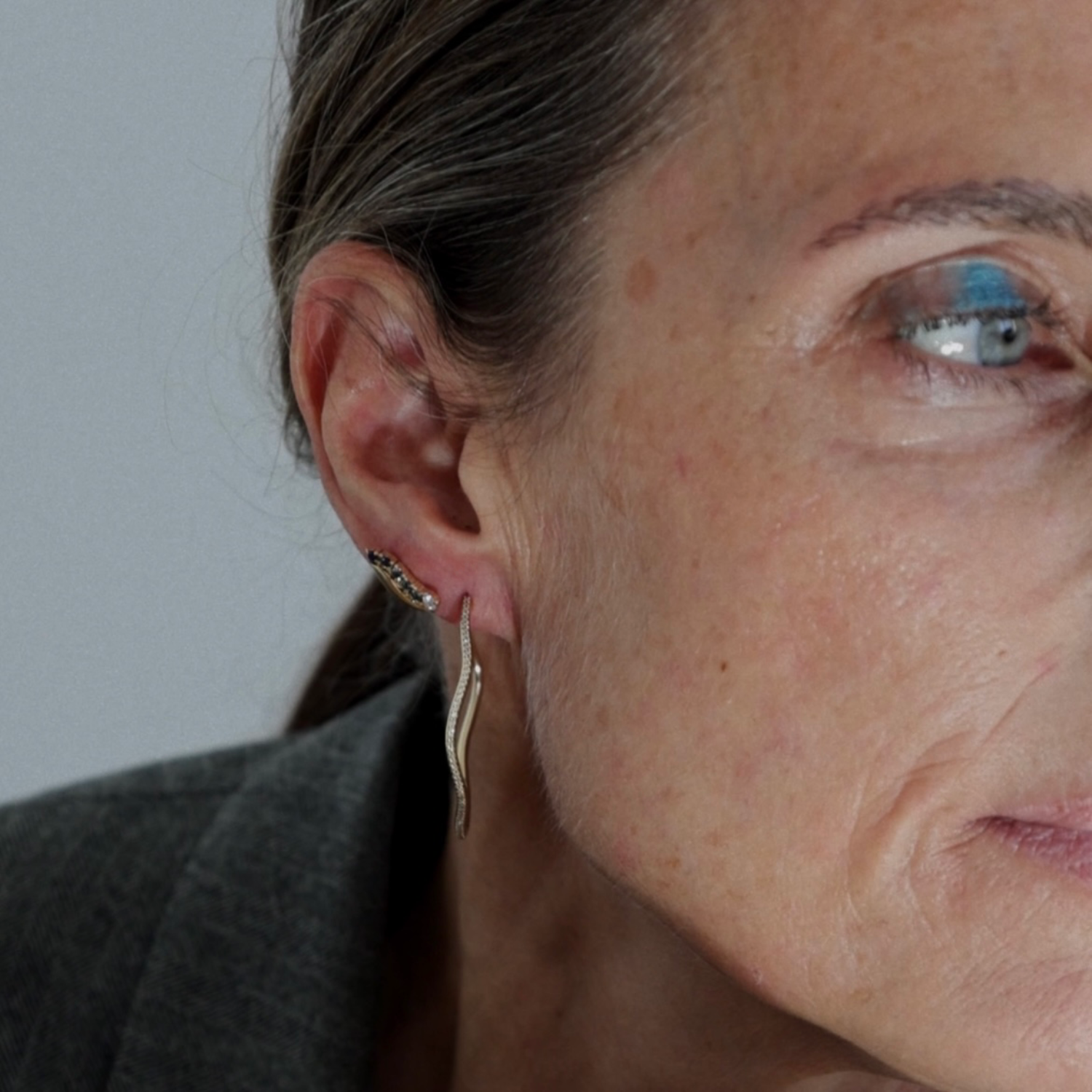 A close-up of a woman's ear wearing a delicate gold huggie hoop earring and a small cluster stud in the lobe, with her face and shoulder visible against a plain light gray background.