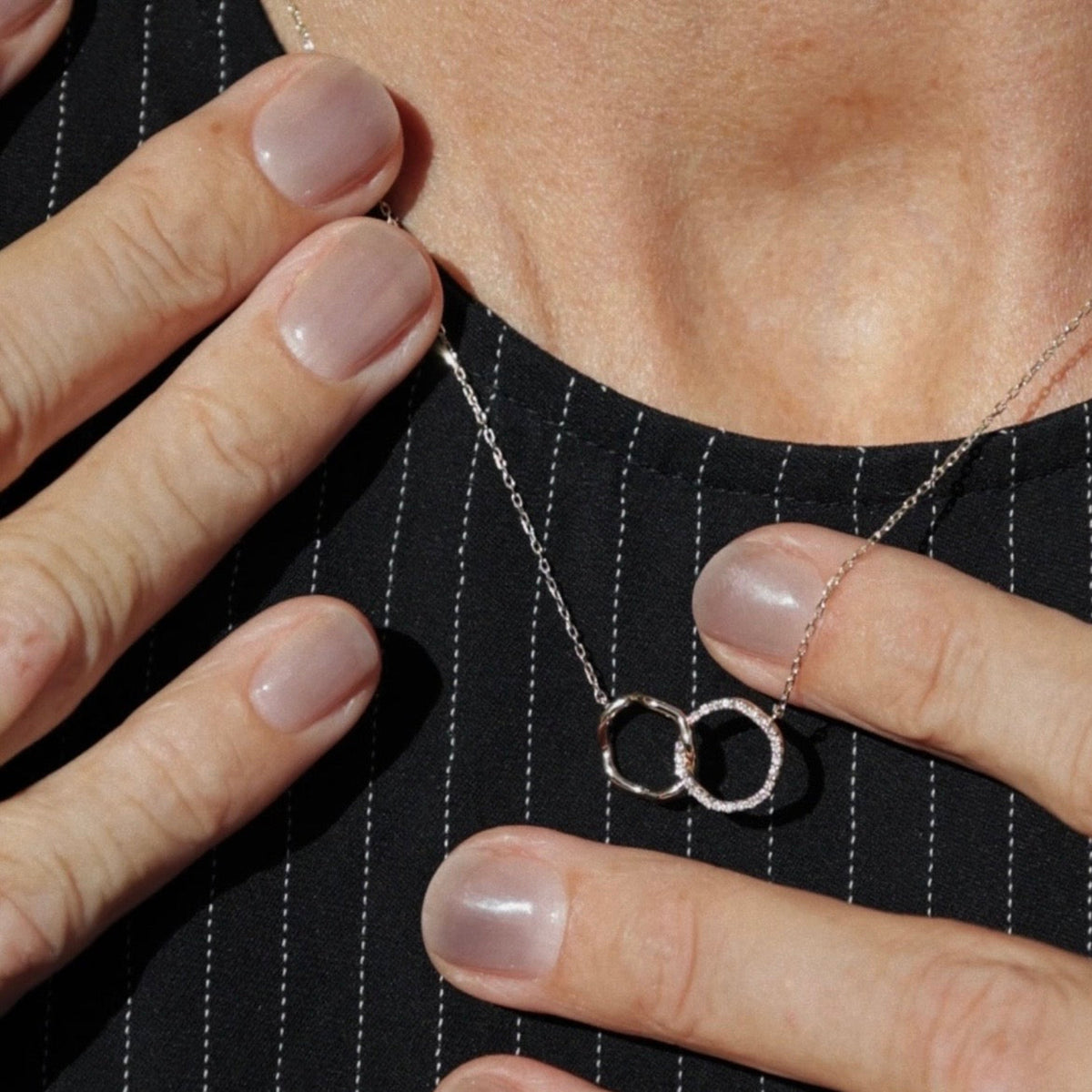 A close-up of a person wearing a delicate silver chain necklace with two interlocking rings pendant, shown against a black pinstriped top.