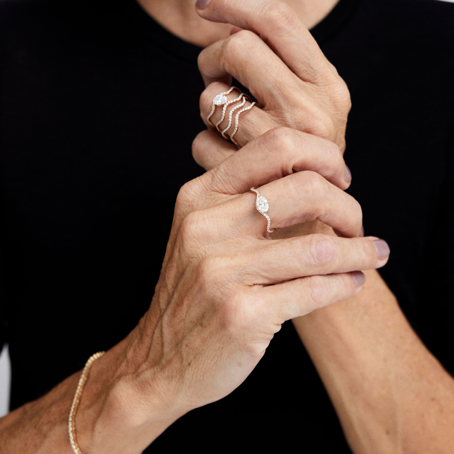 Close-up of a person's hands wearing multiple delicate gold rings, including a stacked wave-style ring and a single ring with a marquise-cut stone, against a black shirt background.