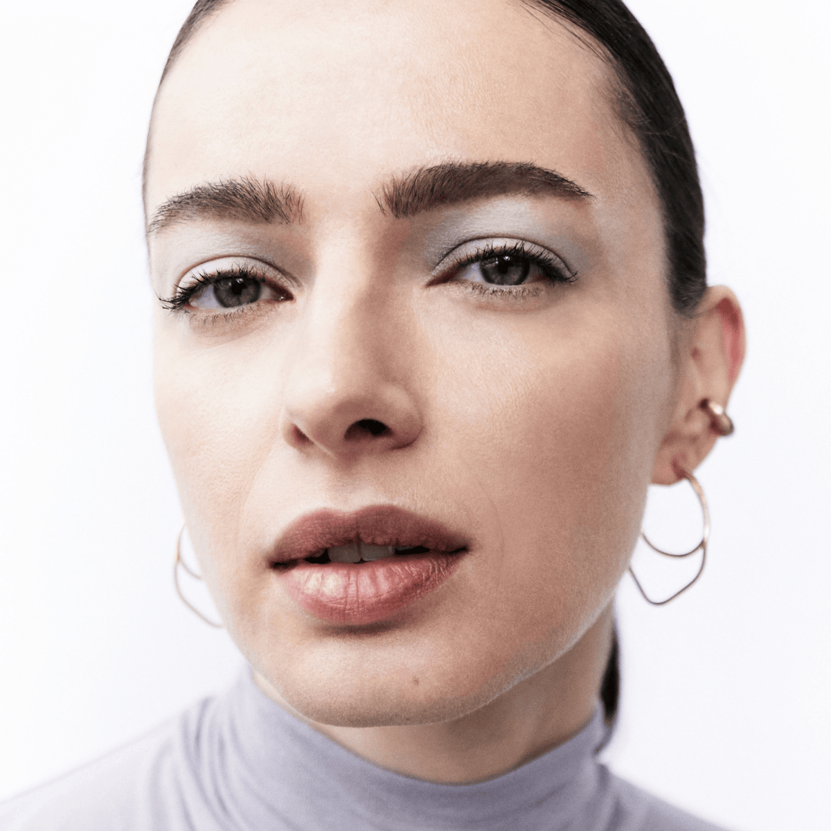 Close-up portrait of a woman wearing large gold hoop earrings and light blue-gray eyeshadow, against a clean white background.