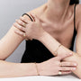 A close-up lifestyle shot of a model wearing a delicate gold jewelry set, including a bangle bracelet, rings, and a ring on the other wrist, against a neutral gray background.