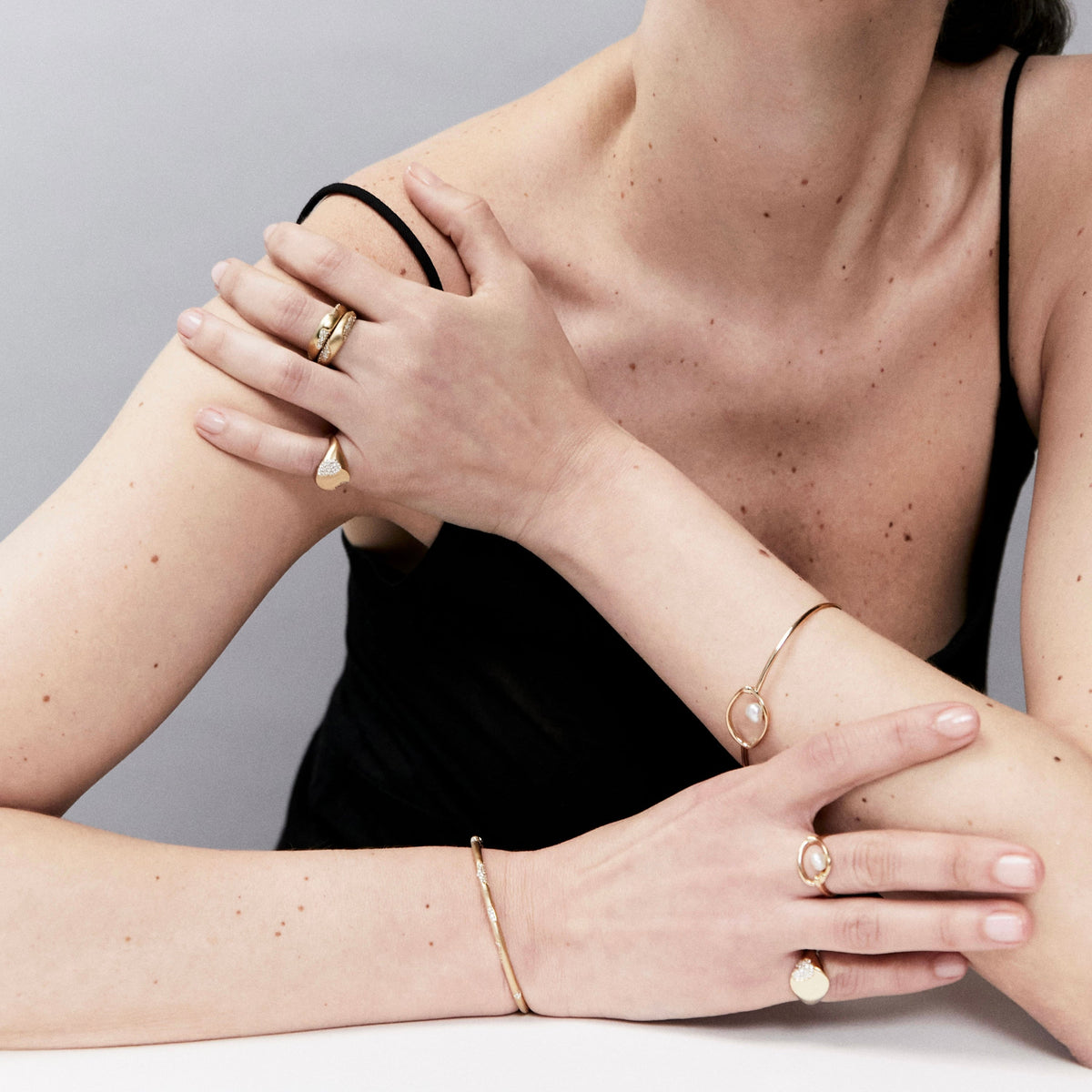 A close-up lifestyle shot of a model wearing a delicate gold jewelry set, including a bangle bracelet, rings, and a ring on the other wrist, against a neutral gray background.