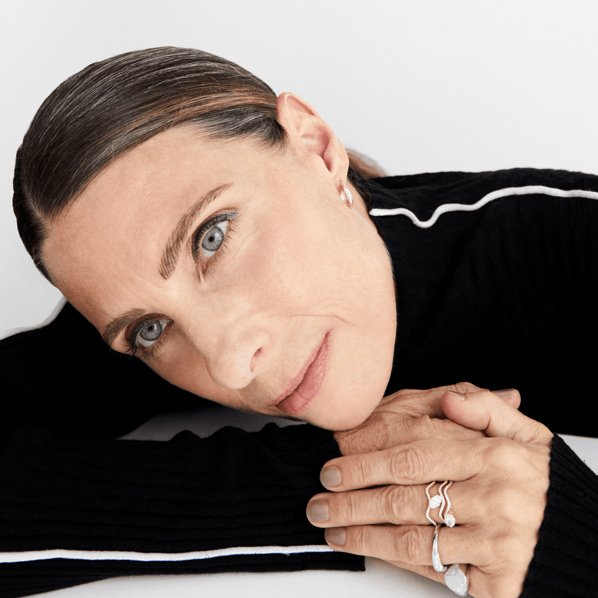 A close-up portrait of a woman with blue eyes resting her head on her hands, wearing silver rings and a small stud earring, dressed in a black ribbed sweater with white stripes against a light gray background.