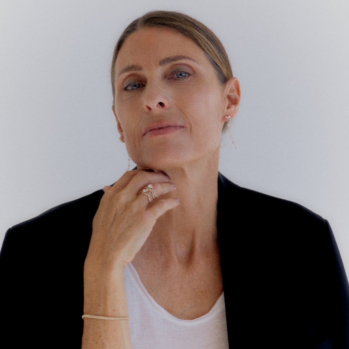 A close-up portrait of a woman with blonde hair pulled back, wearing a black blazer over a white top, with her hand resting near her chin. She is wearing a delicate gold bracelet and ring against a plain light gray background.