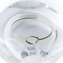 A delicate gold wire bangle bracelet with a small open oval loop, resting on a clear glass dish against a white background.