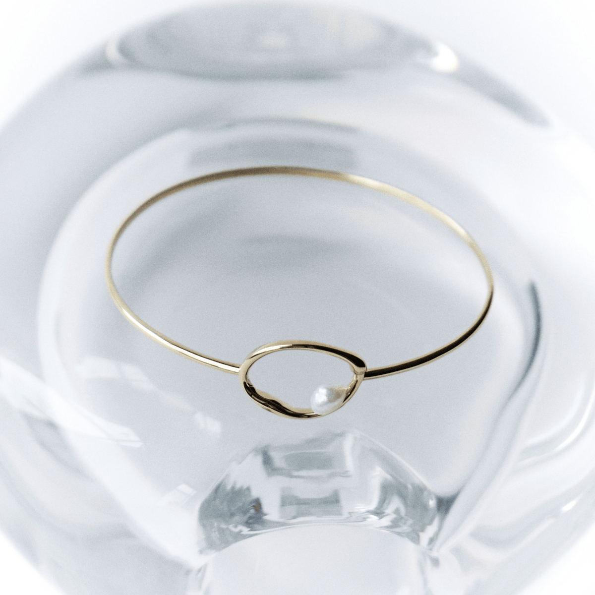 A delicate gold wire bangle bracelet with a small open oval loop, resting on a clear glass dish against a white background.