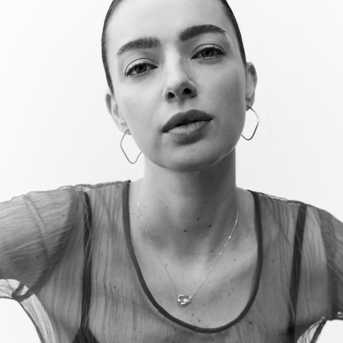 A black and white close-up portrait of a woman wearing large wave hoop earrings, a delicate necklace, and a sheer top against a plain light background.