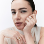 A close-up portrait of a woman wearing multiple gold rings, including one with a large oval clear stone, against a clean white background.