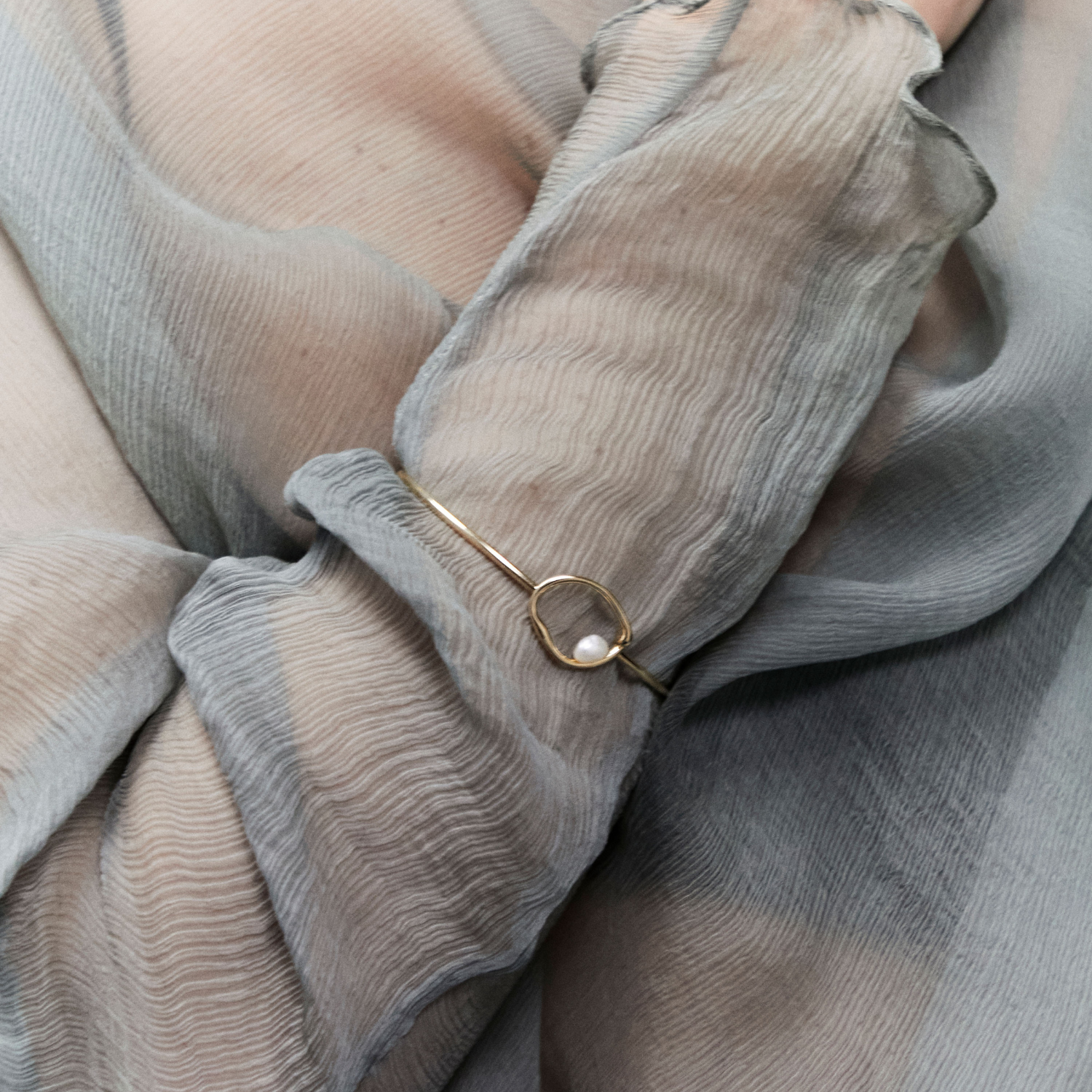 A close-up of a delicate gold ring bangle with a small white pearl, worn on a wrist covered by a sheer, textured gray-blue fabric.