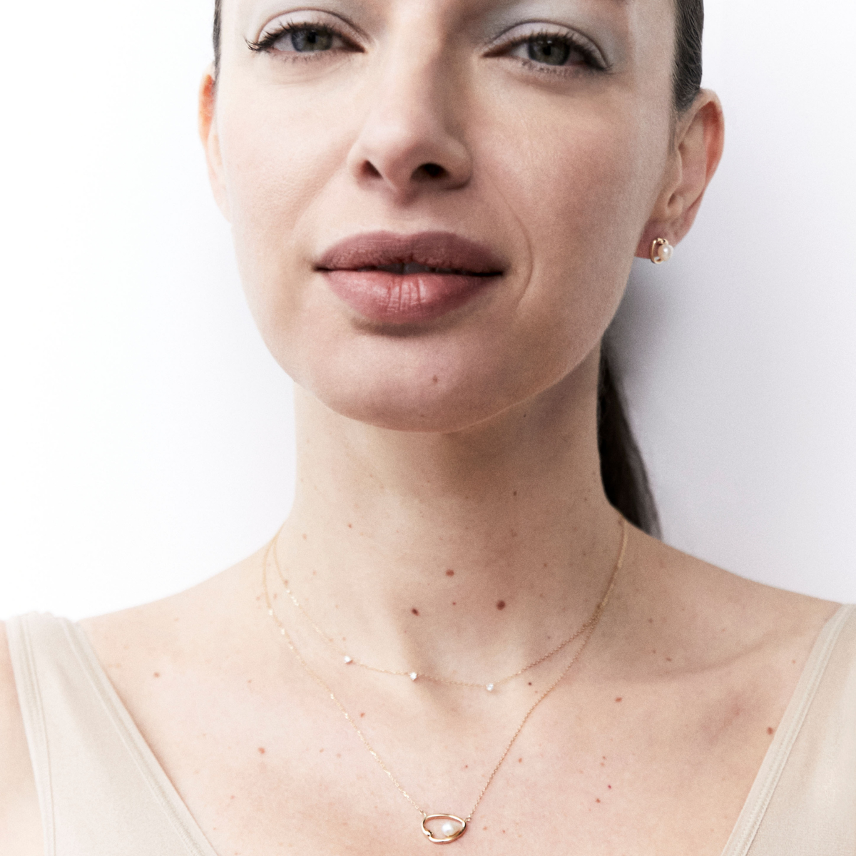 A close-up of a woman wearing a delicate gold double-layer necklace with a small oval pendant, styled with a matching small gold stud earring against a clean white background.
