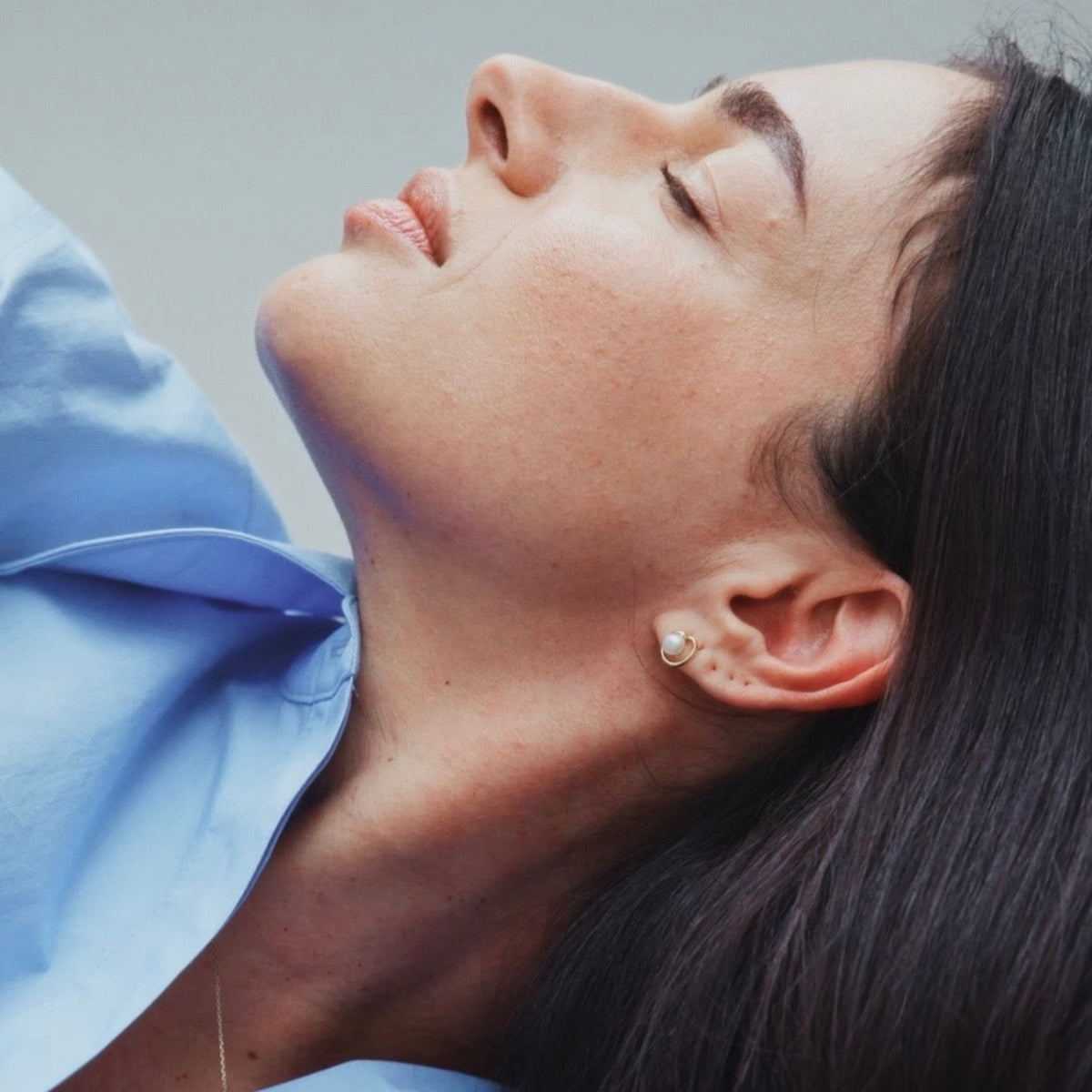 Close-up of a woman with her eyes closed, wearing a small gold hoop earring with a white pearl center and a delicate gold chain necklace, wearing a light blue collared shirt.