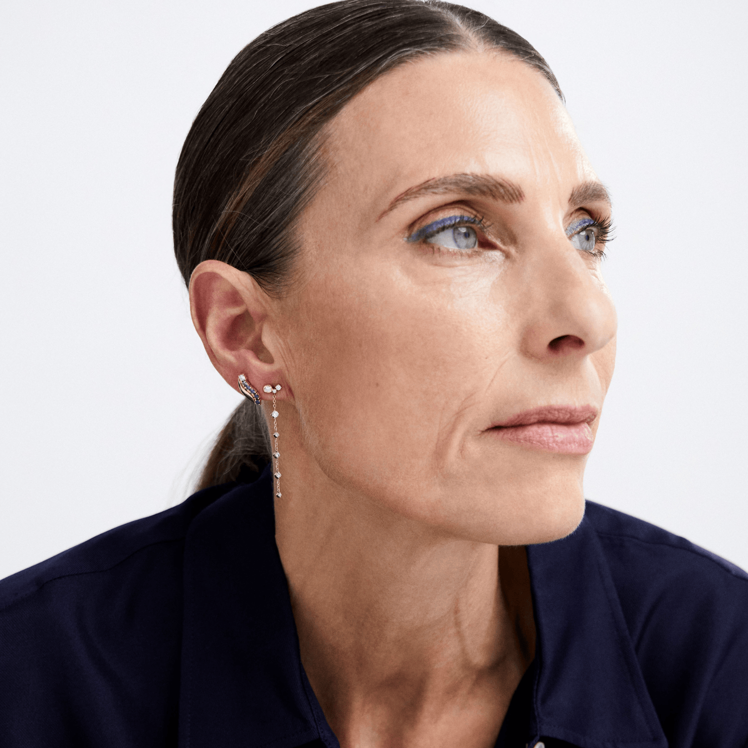 A close-up portrait of a woman wearing a delicate silver dangle earring with small clear stones, set against a clean light gray background.