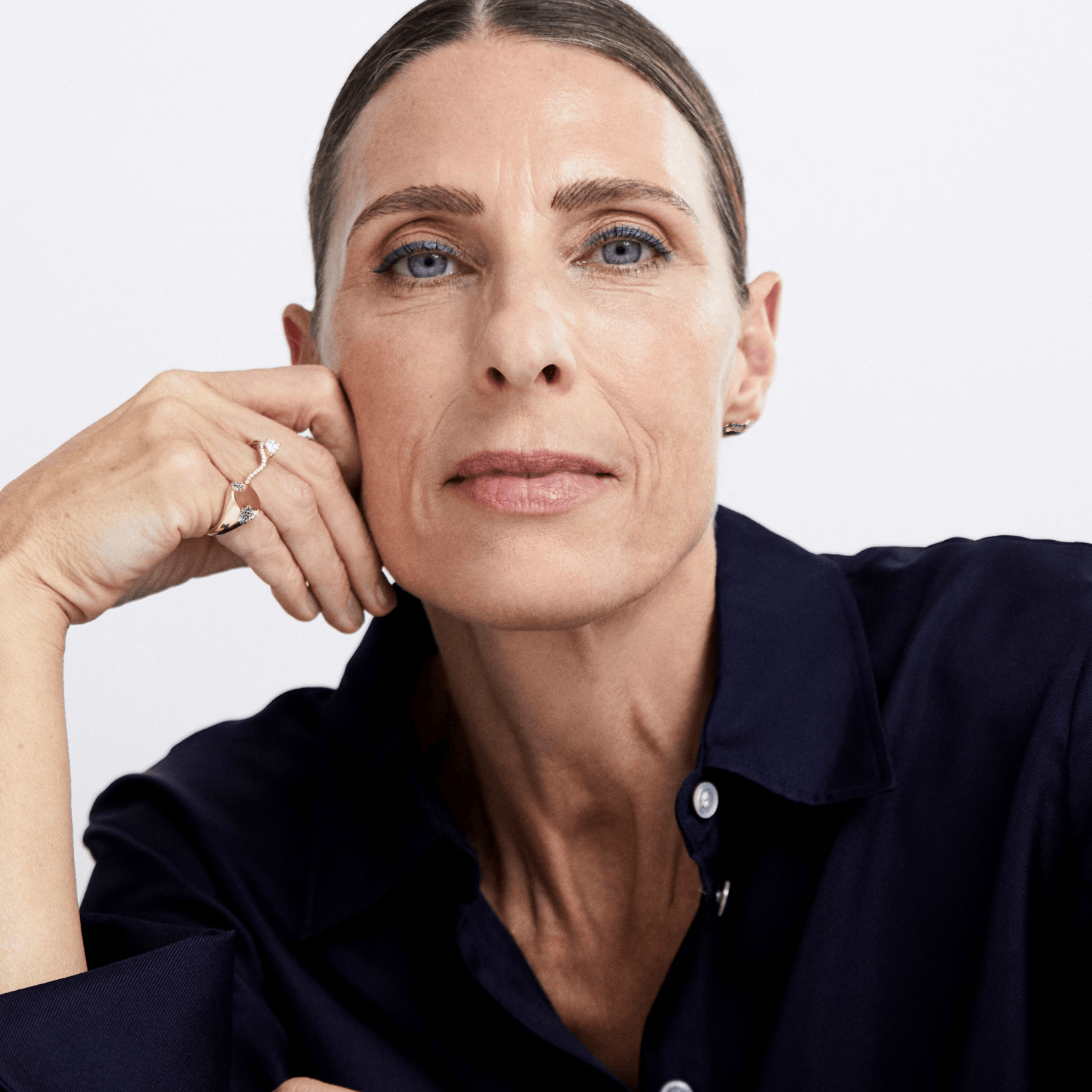 A close-up portrait of a woman with her hand resting on her chin, wearing a dark navy blue button-up shirt and gold rings, set against a plain light gray background.